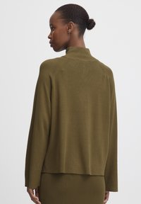 Donna con pelle scura e capelli raccolti in un chignon, che indossa un top a maniche lunghe verde oliva e una gonna a coste abbinata, girata di lato su uno sfondo semplice.