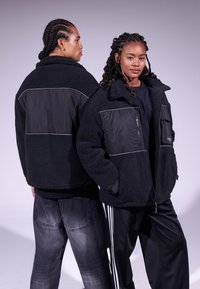 Giacca in pile nera con pannelli a contrasto, chiusura con zip e collo alto. Tessuto liscio, presentata su due modelli fronte e retro.