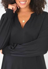 Svart klänning med V-hals, långa ärmar med rynkade detaljer vid ärmsluten och en mjuk tygkvalitet. Guldhalsband med hänge ingår.