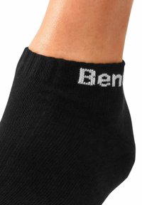 Schwarze Knöchelsocke mit geripptem Muster und kontrastierendem weißen Logo am oberen Rand. Das Material wirkt weich und dehnbar.