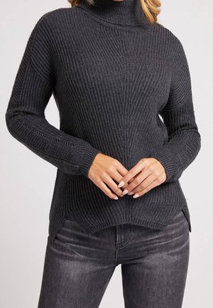 Kvinde iført en mørkegrå ribstrikket rullekravstrøje med lange ærmer og mørkevaske jeans, med hænderne foldet foran taljen.