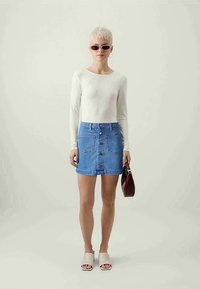 Bela dolga majica z dolgimi rokavi v kombinaciji s svetlo modrim jeans krilom, ki ima sprednje gumbke in žepke. Dodatki vključujejo črne sončne očala in rjavo torbico.