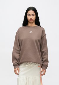 Καφέ φαρδιά φούτερ με στρογγυλή λαιμόκοψη και χαμηλωμένους ώμους, με μικρό λευκό λογότυπο Adidas στο στήθος, σε συνδυασμό με ανοιχτόχρωμη φούστα.