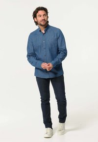 Blauwe jeanshem met lange mouwen, knoopsluiting aan de voorkant, borstzak en een afgeronde zoom, gecombineerd met donkere jeans en witte sneakers.