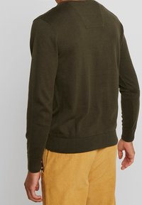 Dunkelgrüner Pullover mit langen Ärmeln, gerippten Bündchen und Saum, mit einem kleinen rechteckigen Patch am hinteren Halsauschnitt. Kombiniert mit Cordhosen.