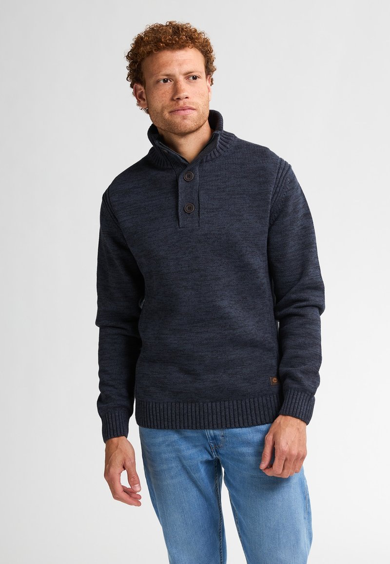 Bărbat cu păr creț, îmbrăcat într-un pulover tricotat încheiat cu nasturi de culoare albastru închis și blugi de culoare albastru deschis, stând pe un fundal alb simplu.