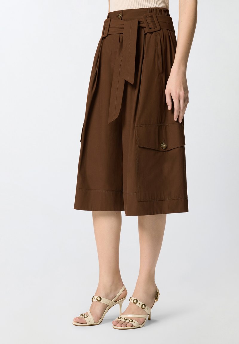 Καφέ cargo σορτς με ψηλή μέση και φαρδιά μπατζάκια, με ζώνη και τσέπες με κουμπιά, φορεμένα με μπεζ σανδάλια με τακούνι.