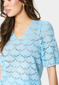 Blusa blu in pizzo con scollatura a V, maniche a sbuffo e motivo a onde, che mette in evidenza un materiale trasparente e testurizzato.