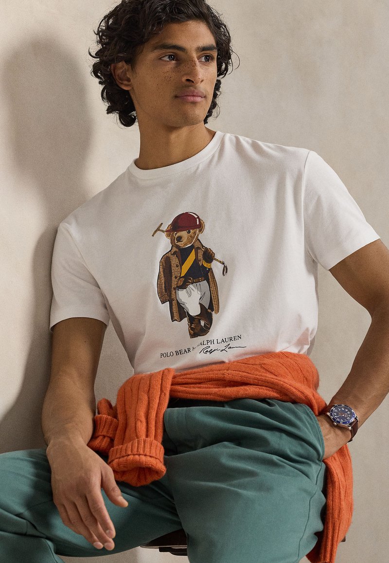 Weißes Baumwoll-T-Shirt mit einer Grafik eines Bären in einem braunen Karo-Mantel. Das Modell trägt türkise Hosen und einen orangen Strickpullover um die Taille gebunden.