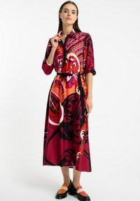 Kobieta w midi sukience z długim rękawem i paskiem, w odważne, abstrakcyjne wzory w kolorze czerwonym, pomarańczowym i czarnym, w połączeniu z mokasynami w kolorze blokowym.