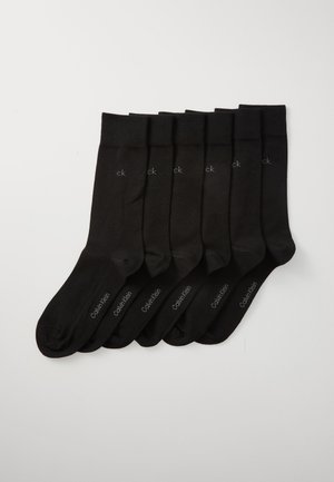 Seis pares de calcetines negros de Calvin Klein alineados ordenadamente sobre un fondo blanco, cada uno con "ck" cerca del puño y "Calvin Klein" en el pie.