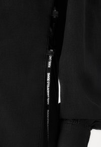Juodas švarkas su lygiu audiniu, užtrauktuko detale ir etikete su išdidžiai i�šspausdintu "KARL LAGERFELD JEANS". Papuošti rankogaliai.