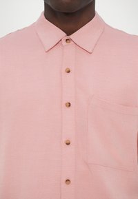 Light pink short-sleeve shirt made of textured fabric, featuring a buttoned front, wooden buttons, and a patch pocket on the left side.