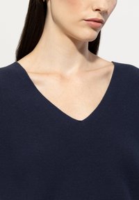 Dunkelblauer Strickpullover mit tiefem V-Ausschnitt, sichtbarer Textur und dezentem Rippenmuster an den Schultern. Nahaufnahme von Ausschnitt und Schlüsselbein.