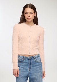Cardigan corto fluffy di un delicato rosa chiaro, con maniche lunghe e scollo rotondo. Presenta sei bottoni simili a cristalli lungo la parte anteriore.