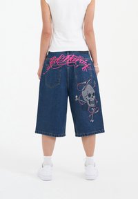 Denim shorts with a loose fit, featuring a pink embroidered logo and a skull design with accents. Standard back pockets and cuffed hems.