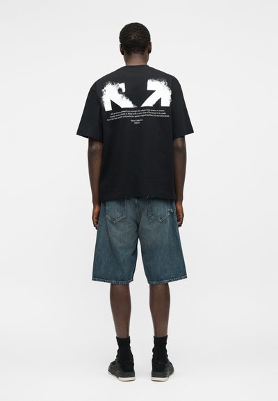 Άνδρας που φοράει μαύρο φαρδύ μπλουζάκι με λευκά βέλη και κείμενο στην πλάτη, μπλε τζιν σορτς, μαύρες κάλτσες και μαύρα αθλητικά παπούτσια, γυρισμένος προς τα πίσω.