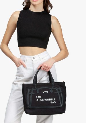 Donna che indossa un top corto nero senza maniche e pantaloni bianchi, che tiene una borsa a mano nera con la scritta bianca "SONO UNA BORSA RESPONSABILE."