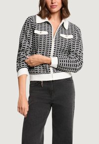 Cardigan a fantasia bianco e nero con colletto bianco, chiusura con zip e due tasche frontali. Il tessuto appare strutturato e aderente.
