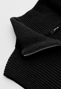 Nahaufnahme von schwarzem geripptem Strickstoff mit einem teilweise offenen silbernen Reißverschluss und einem kleinen schwarzen Etikett mit der Aufschrift "OPUS."