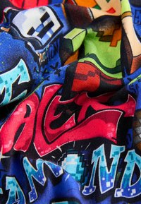 Bunt gemusteter Stoff mit einer Mischung aus grafischen Designs in Blau, Rot, Grün und Schwarz. Text- und Cartoon-Charaktermuster in einem weichen, strukturierten Gewebe.