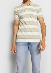 Béžové a bílé pruhované polo tričko s klasickým límečkem, krátkými rukávy a malým červeným logem na hrudi, doplněné světle modrými džínami.