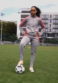 Sportovní tepláková souprava v šedé barvě s červenými akcenty; těsně padnoucí bunda a kalhoty. Stojí na černo-bílém fotbalovém míči na zeleném trávníku.
