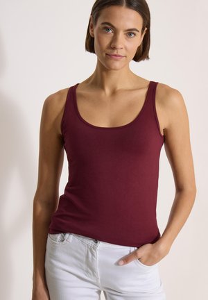 Femme aux cheveux courts et bruns portant un haut bordeaux sans manches et un pantalon blanc, debout avec une main dans la poche devant un fond uni.
