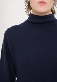 Personne portant un pull à col roulé côtelé bleu marine foncé, avec des cheveux courts et bouclés et des lèvres neutres sur un fond clair.