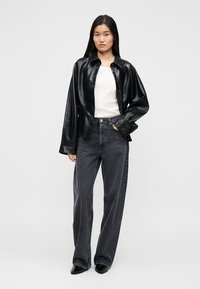 Μαύρο πουκάμισο από ψεύτικο δέρμα σε υπερμεγέθη γραμμή, λευκό εφαρμοστό τοπ και σκούρο γκρι τζιν ψηλόμεσο. Το μοντέλο φοράει μαύρα μυτερά παπούτσια και στέκεται με τα χέρια στις τσέπες.