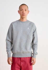Felpa grigia con scollo rotondo e polsini a coste. Presenta un piccolo logo bianco sul petto. Indossata con pantaloni rossi.