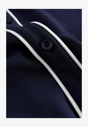 Marineblåt stof med en glat tekstur har hvide kanter og en enkelt sort knap. Designet fremhæver enkelhed og elegance.