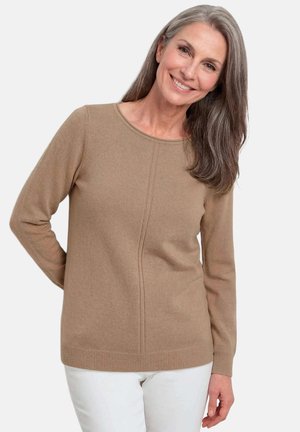Lachende oudere vrouw met lang grijs haar, een beige trui en witte broek, staand tegen een effen lichte achtergrond.