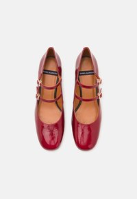 Red patent leather Mary Jane shoes with a square toe, two strap buckles, and an open heel. Smooth surface with a tan inner lining.