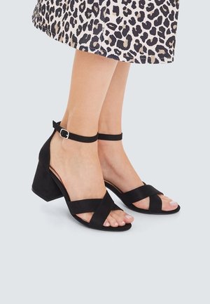 Pieds portant des sandales noires à talons blocs et à brides, associées à une jupe à imprimé léopard sur un fond neutre.