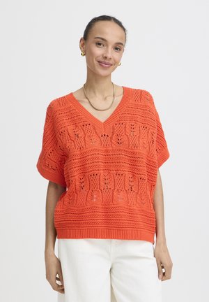 Nasmejana žena s zlatnim obručima za uši i ogrlicom, narančastom pletenom bluzom s V-izrezom i bijelim hlačama na jednostavnoj svijetloj pozadini.