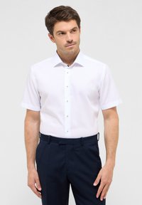 Camicia bianca a maniche corte con colletto a bottoni, caratterizzata da un bordo del colletto in contrasto e bottoni argentati, abbinata a pantaloni blu scuro.