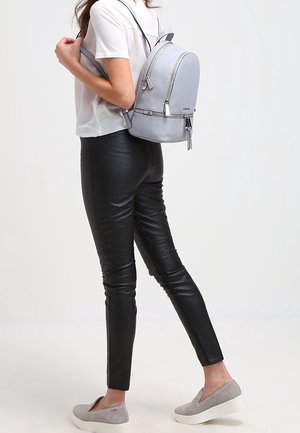 Mochila de cuero gris claro con forma redondeada, bolsillo frontal con cremallera y correas ajustables, usada sobre una camiseta blanca y pantalones negros ajustados.