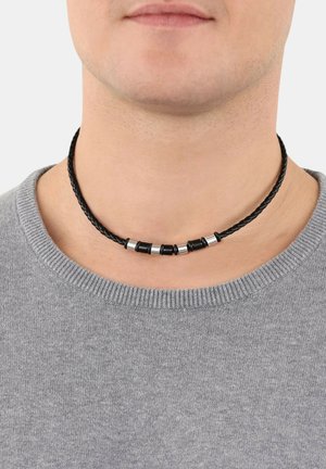 Collana choker in pelle nera intrecciata con accenti in metallo, argentati e neri, indossata attorno al collo, sopra un maglione grigio con scollatura tonda.