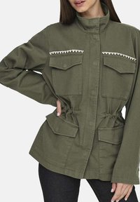 Veste utilitaire vert olive avec deux poches à rabat, taille cintrée et décorations en strass sur la poitrine. Manches longues.