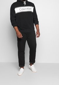 Felpa nera in cotone con logo bianco "Calvin Klein" e strisce. Abbinata a pantaloni neri e sneakers bianche. Tessuto morbido, vestibilità rilassata.