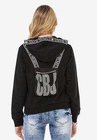 Black hoodie featuring a patterned hood, adjustable straps with text, and a rhinestone-embellished pocket on the back.