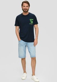 Marineblauwe katoenen t-shirt met groene grafische bedrukking op de borst, gecombineerd met lichtblauwe denim shorts en witte sneakers. Casual fit, korte mouwen.