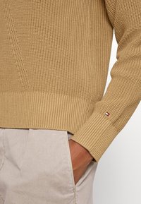 Maglione lavorato a maglia beige con texture, con polsini e orlo a coste. Presenta un piccolo logo multicolore vicino al polso. Design morbido e casual.