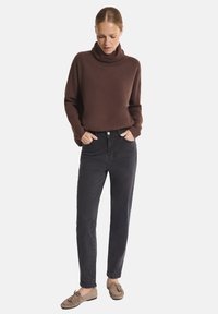 Kobieta w brązowym swetrze z golfem, czarnych jeansach o wysokim stanie i beżowych mokasynach stoi z rękami w kieszeniach.