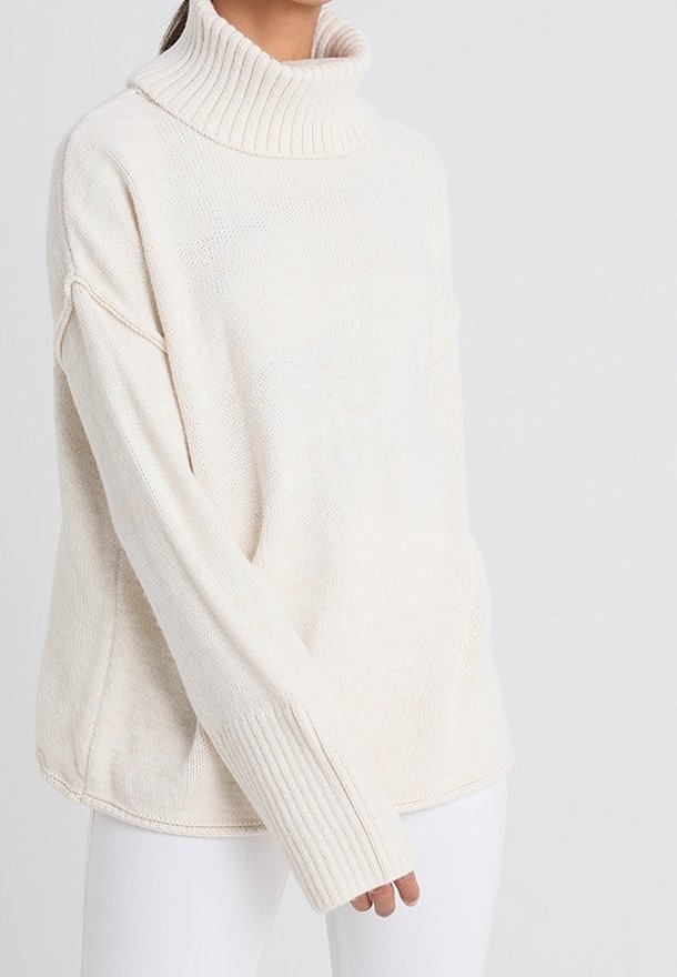 Femme portant un pull col roulé ample crème avec de longues manches côtelées et un pantalon blanc sur fond uni.
