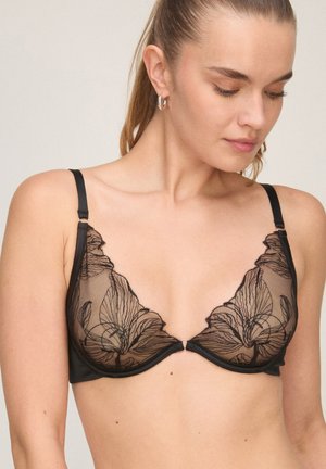 Femme portant un soutien-gorge noir transparent avec broderie florale, regardant vers le bas, cheveux attachés et boucles d'oreilles créoles argentées.