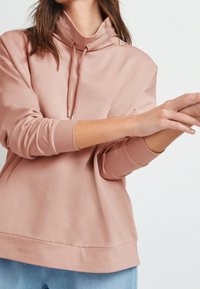 Una persona con una sudadera rosa claro de cuello alto está moviendo las manos, con un fondo blanco simple.