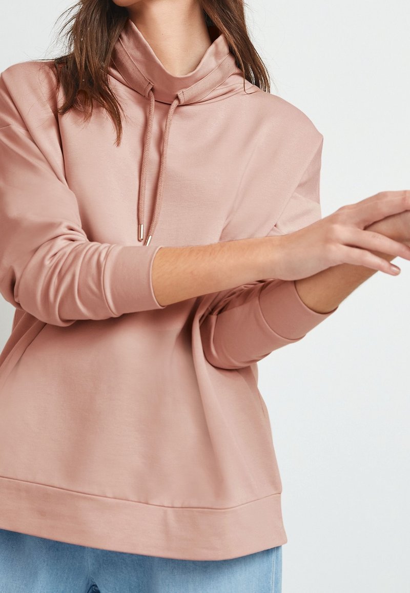 Una persona con una sudadera rosa claro de cuello alto está moviendo las manos, con un fondo blanco simple.