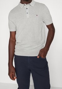 Grå stickad polo med krage, försedda med tre knappar och en liten logotyp på vänster bröst, kombinerad med mörkblå byxor.